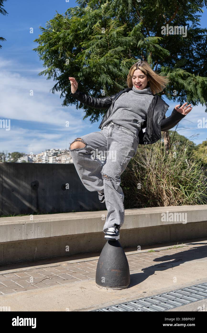 La giovane donna alla moda si bilancia con sicurezza su un paletto, mostrando il suo stile unico e il suo spirito spensierato in un ambiente urbano contemporaneo Foto Stock