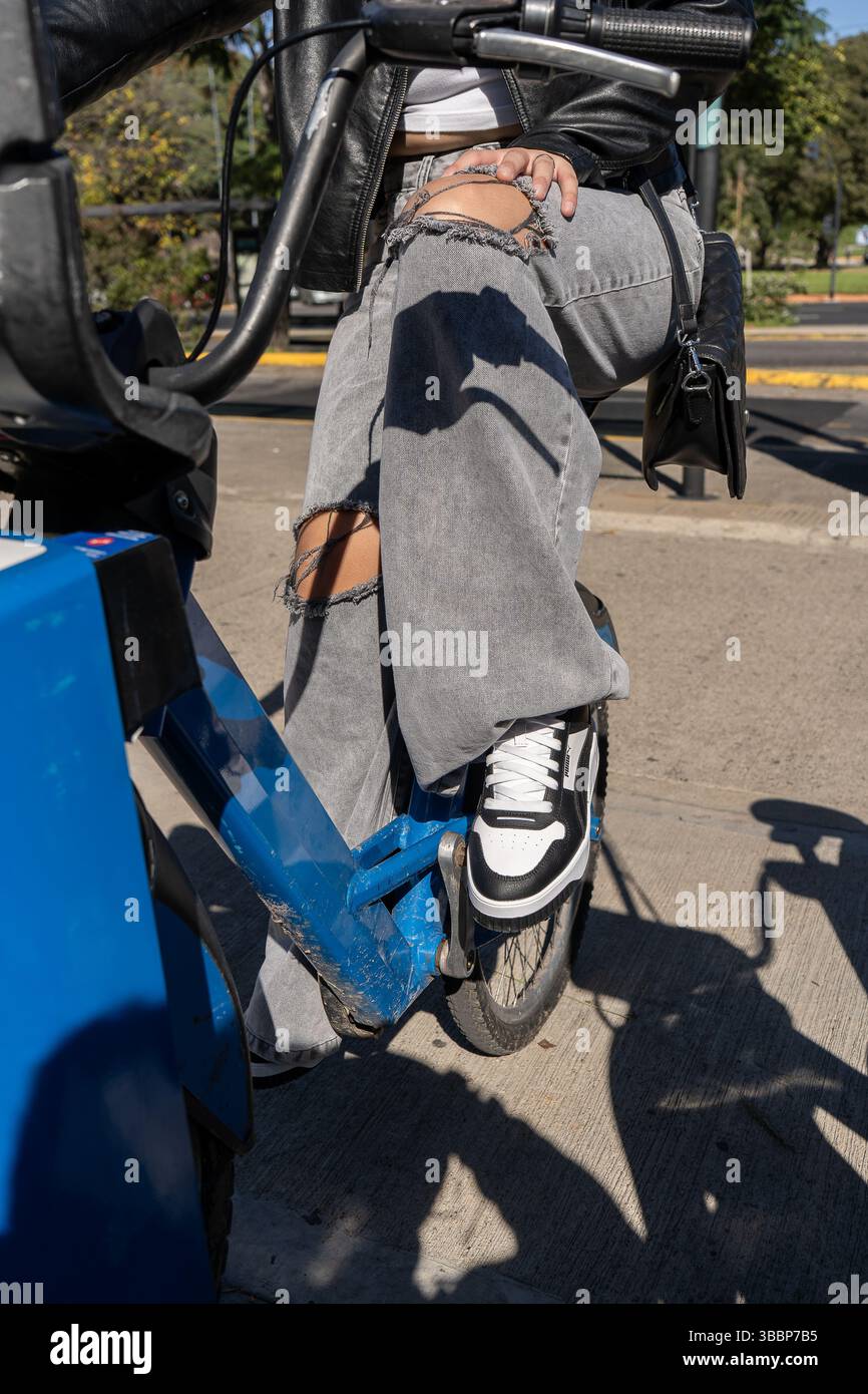 Primo piano delle gambe di una giovane donna in jeans strappati e sneakers, seduti su una bici a noleggio blu, godendosi una giornata di sole in città Foto Stock
