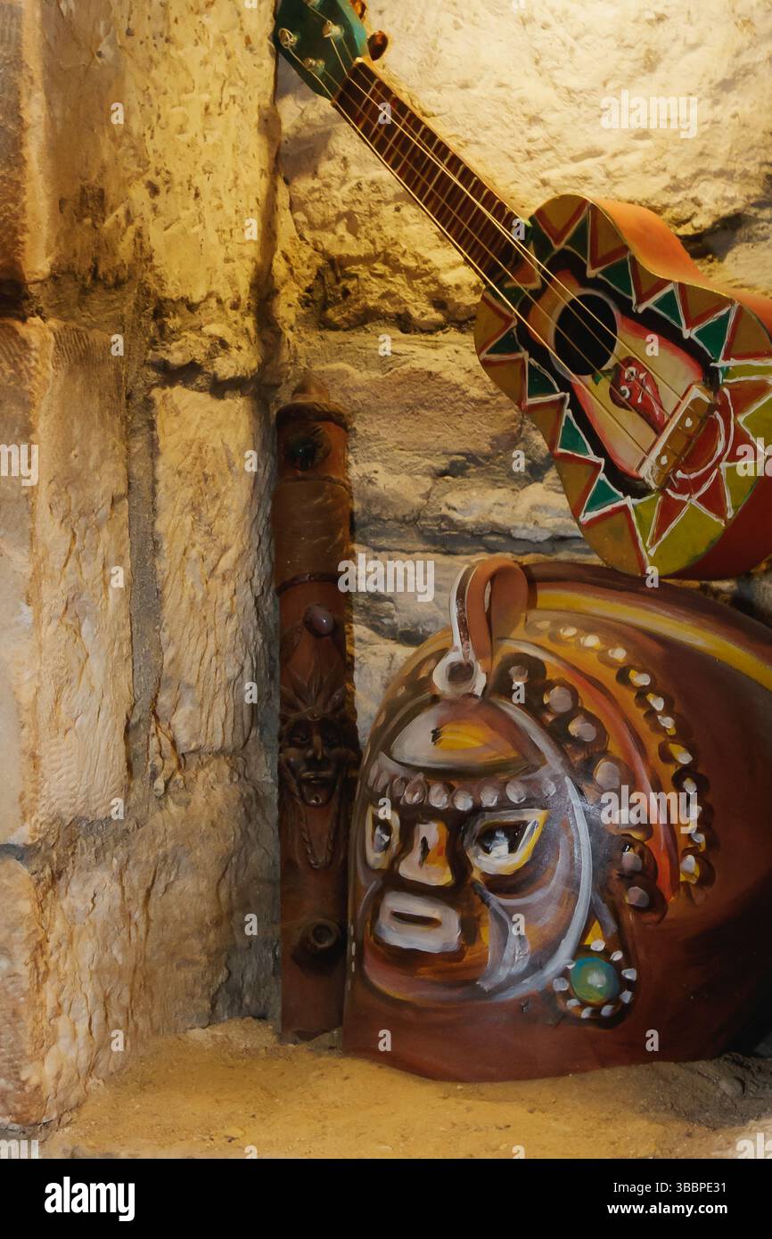 Una vibrante collezione di arte popolare è disposta su una mensola in pietra, con una chitarra dipinta, maschere tradizionali e figure in legno intagliato, che mostrano cu Foto Stock