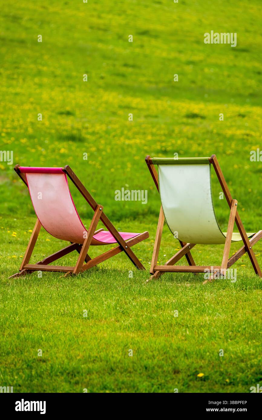 Due sedie a sdraio in legno con imbracature di tela pastello disposte su un pendio erboso verdeggiante sotto la luce diffusa del giorno Foto Stock