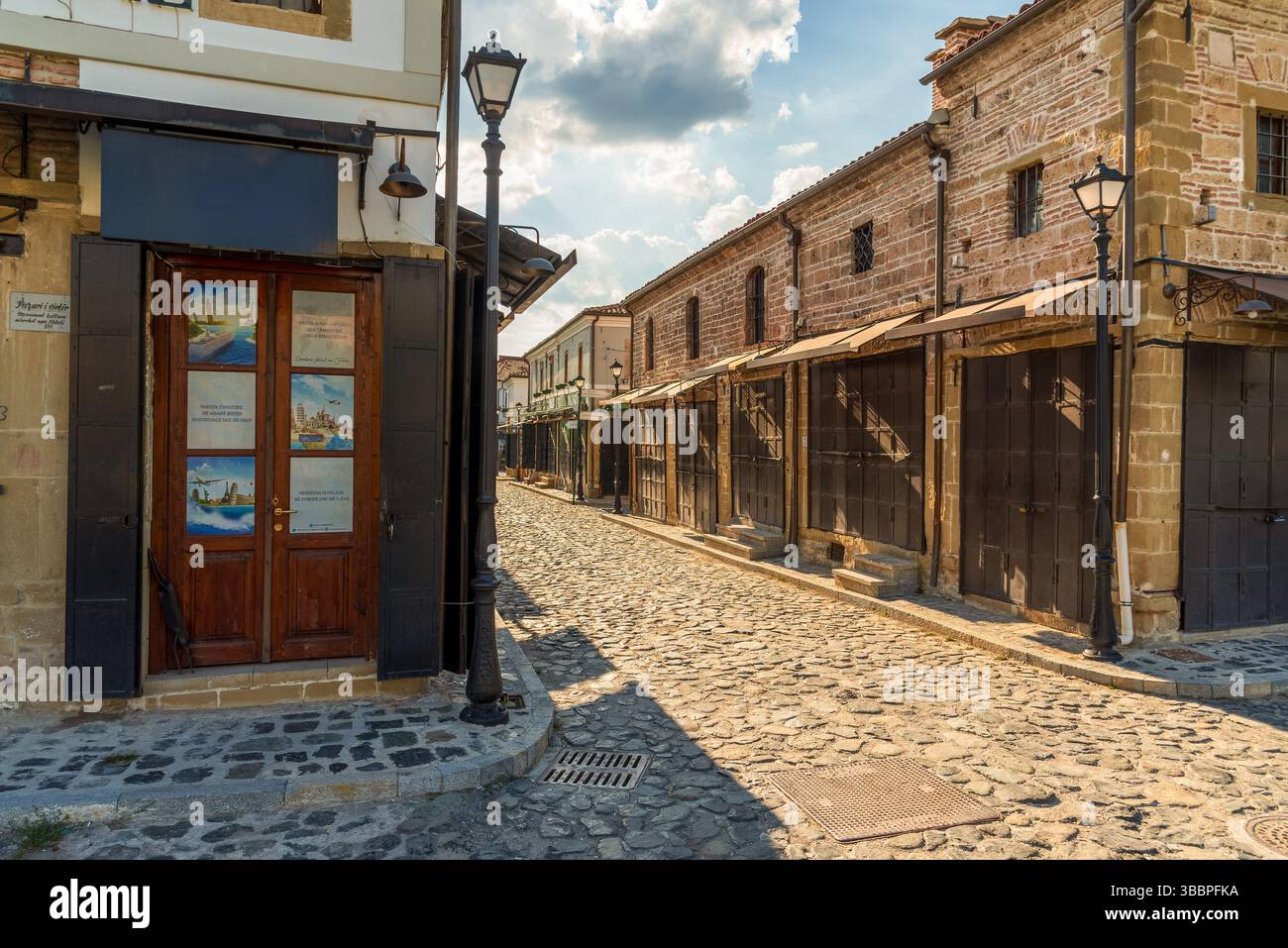 Vicolo acciottolato a Korca, Albania, fiancheggiato da negozi con facciata in pietra e lampioni ornati in ferro battuto sotto un cielo estivo cristallino Foto Stock