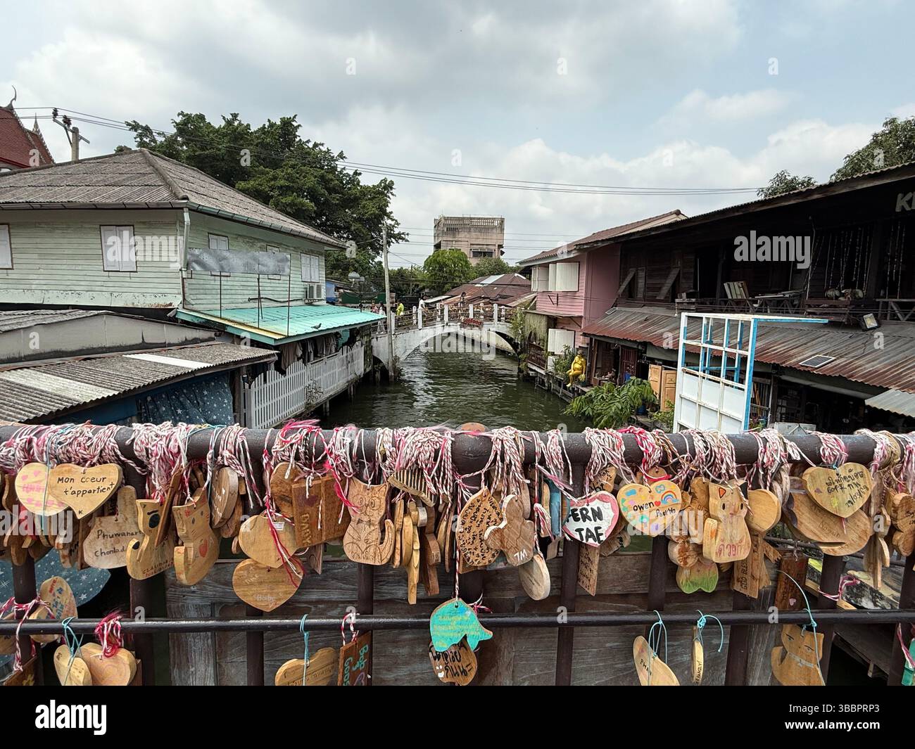 Targhe a forma di cuore appese su un ponte sul canale in Thailandia, simboleggiano l'amore e la connessione nel tranquillo ritmo della vita fluviale locale - Immagine stock catturata con smartphone