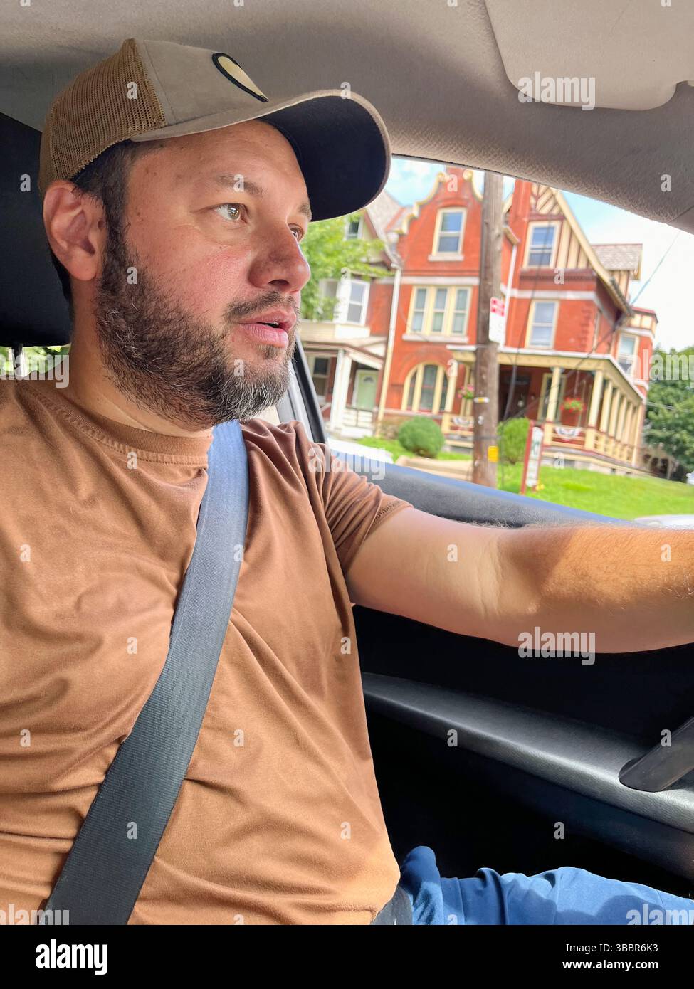 Un uomo che indossa una camicia marrone e un cappello sta guidando un'auto. Sta indossando una cintura di sicurezza Foto Stock Un uomo che indossa una camicia marrone e un cappello sta guidando un'auto. Sta indossando una cintura di sicurezza Foto Stock