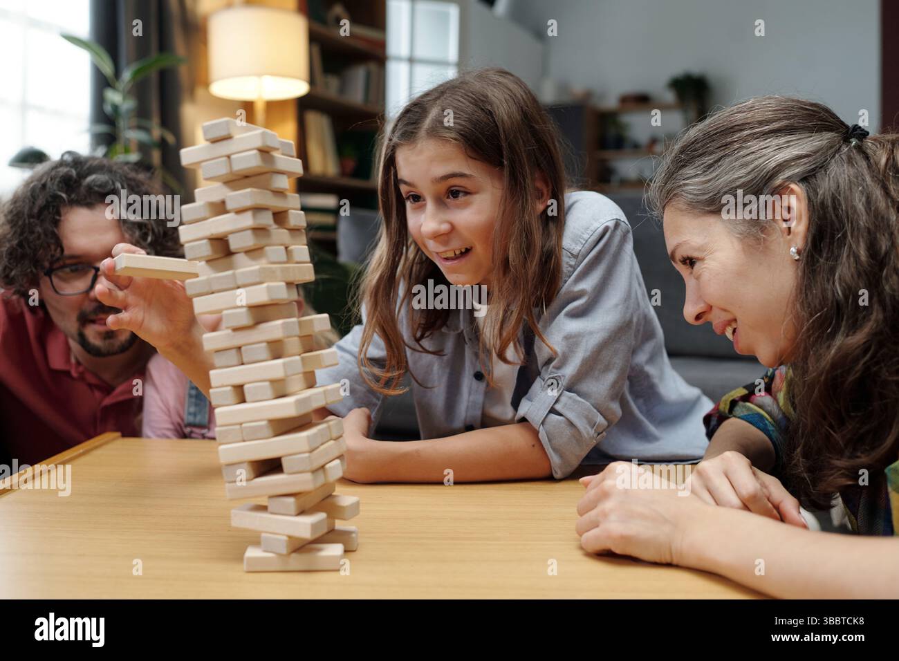 I membri della famiglia si divertono insieme mentre costruiscono un'alta torre utilizzando blocchi di legno nel soggiorno. Sorrisi e risate creano un'atmosfera gioiosa e coinvolgente Foto Stock