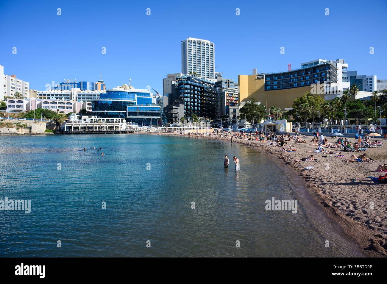 Spiaggia nella baia di St George con turisti in una bella giornata di sole e hotel moderni sullo sfondo, St Julian's, Malta. Foto Stock
