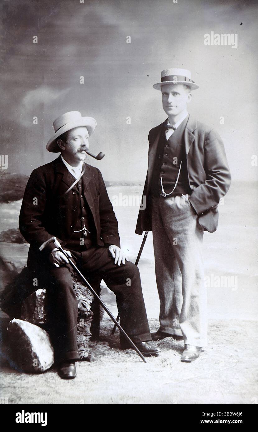 Storia sociale, moda: Ritratto di due gentiluomini ben vestiti, ognuno con canne e cappelli (diportisti). L'uomo seduto sta fumando una pipa, il suo collega sta in piedi con una mano in tasca. Non attribuito, intorno al 1910. Da una collezione mista di cartoline edoardiane. Foto Stock