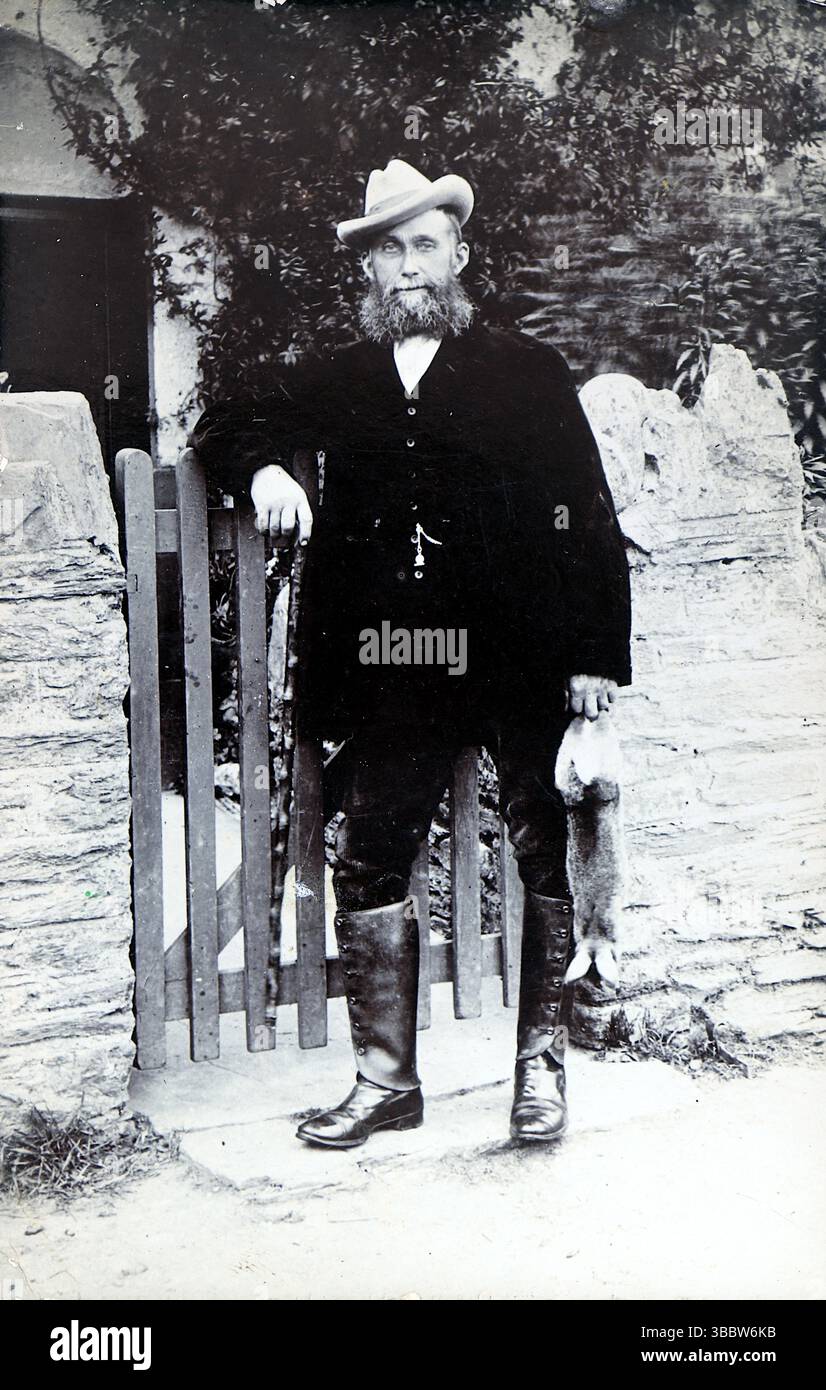 Storia sociale, moda: Un gentiluomo in piedi accanto a un cancello in un ambiente rurale. L'uomo indossa abiti neri, un gilet, stivali al ginocchio e un cappello. Ha una barba grande e tiene una lepre o un coniglio nella mano sinistra. Non attribuito, pubblicato intorno al 1908. Da una collezione mista di cartoline edoardiane. Foto Stock