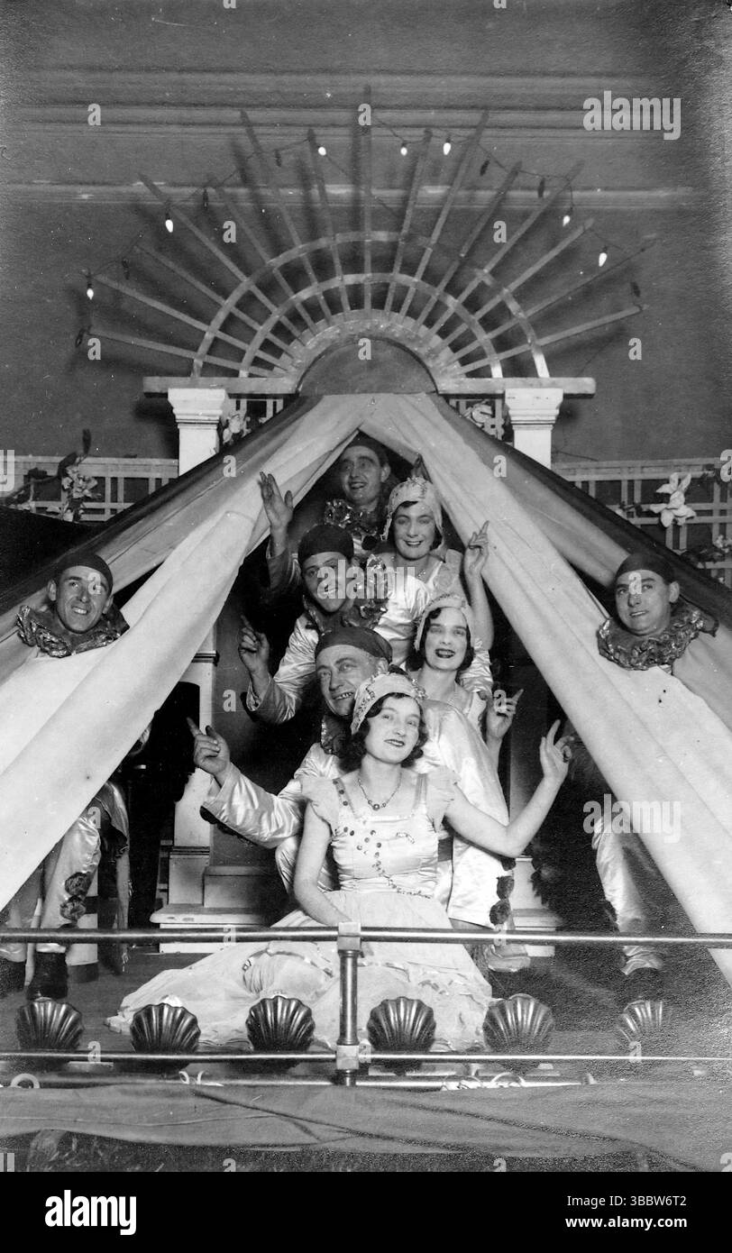 Un gruppo di persone, animatori, ha posato all'interno di un palcoscenico che assomiglia a una grande tenda o tenda da sole. In primo piano, una donna con un vestito di colore chiaro è seduta, con diversi altri individui che guardano da dietro e sopra di lei, alcuni con trucco teatrale o costumi, tra cui quelli che sembrano essere colletti da clown su due figure che sbirciano dai lati. Sullo sfondo, c'è una struttura decorativa che ricorda un ventilatore o un'esplosione di sole. L'impressione generale è di uno spettacolo teatrale. Da una collezione mista di cartoline edoardiane. Foto Stock