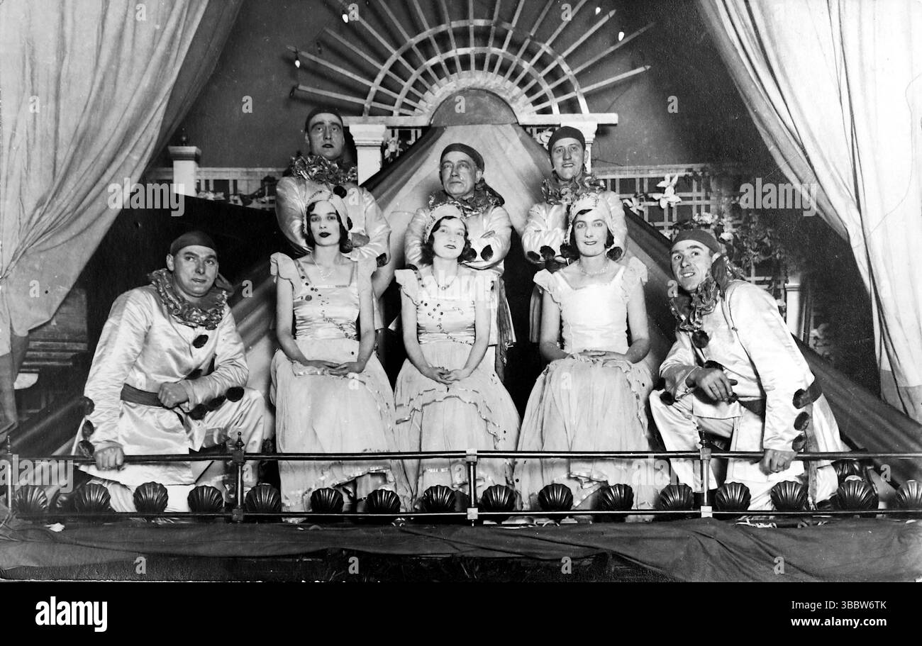 L'immagine mostra un gruppo di persone, possibilmente intrattenitori, poste all'interno di quello che sembra un palcoscenico simile a una grande tenda o tenda da sole. In primo piano, una donna con un vestito di colore chiaro è seduta, con diversi altri individui che guardano da dietro e sopra di lei, alcuni con trucco teatrale o costumi, tra cui quelli che sembrano essere colletti da clown su due figure che sbirciano dai lati. Sullo sfondo, c'è una struttura decorativa che ricorda un ventilatore o un'esplosione di sole. Da una collezione mista di cartoline edoardiane. Foto Stock