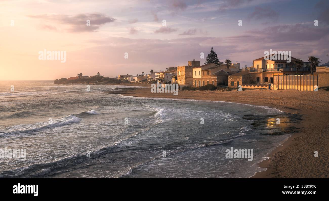 Le onde si infrangono dolcemente contro la spiaggia sabbiosa mentre il sole tramonta dietro le affascinanti case di Punta secca, in Sicilia, offrendo pace, in Italia Foto Stock