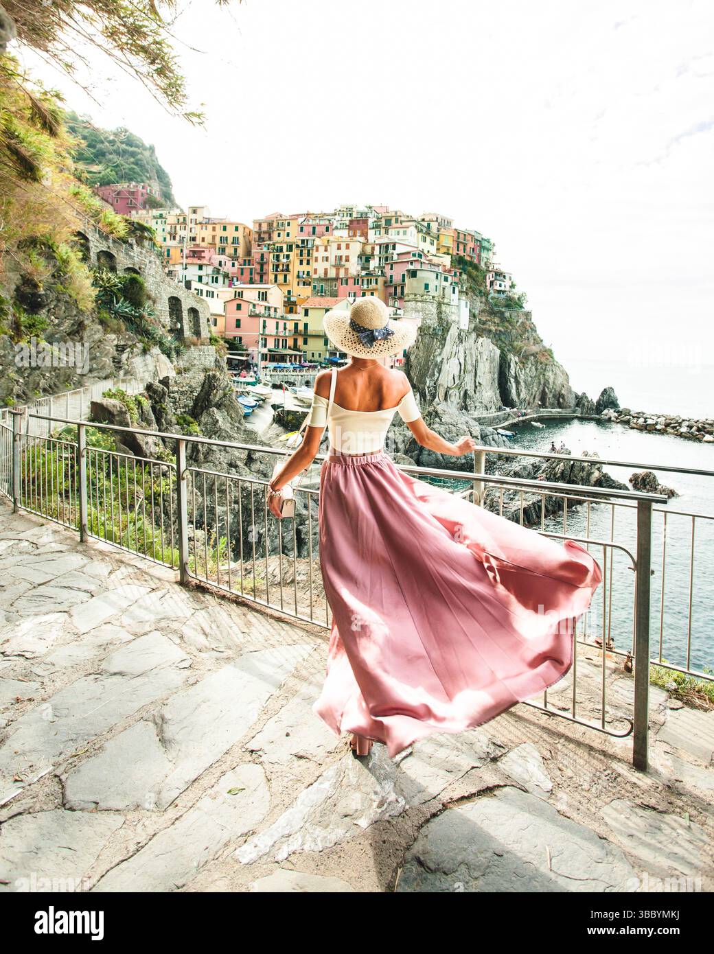 La giovane donna alla moda si sta allontanando dalla macchina fotografica e guarda la vista panoramica delle cinque Terre di Manarola, in Italia, mentre la sua lunga gonna in seta rosa Foto Stock