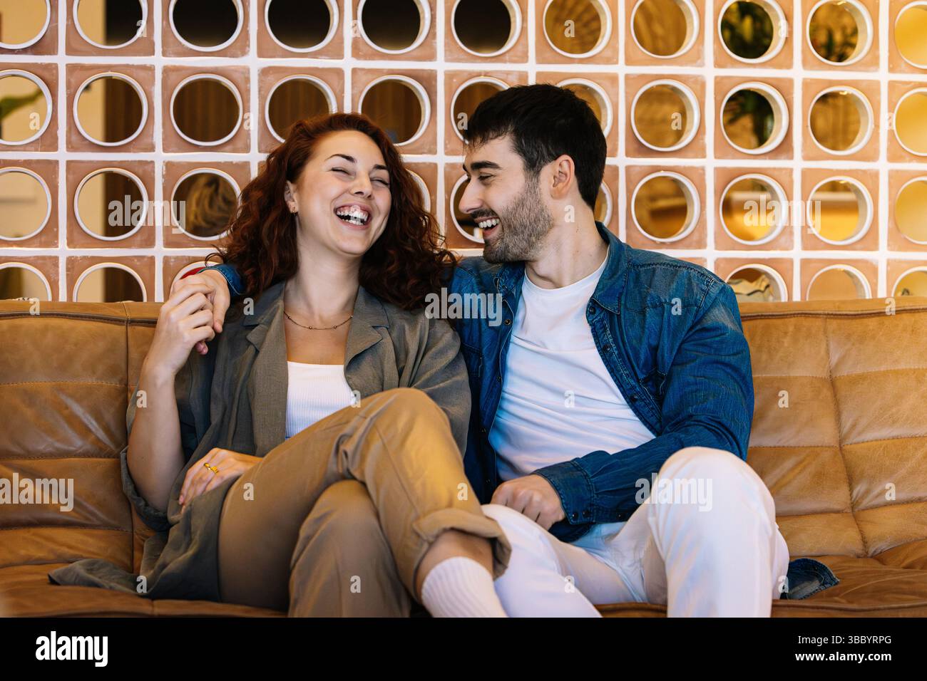 Una giovane coppia gioiosa si diverte a ridere mentre si rilassa su un divano in pelle abbronzata in una lounge interna decorata con calore e con elementi di design moderni. Vite umane Foto Stock