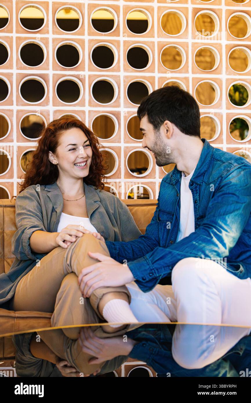 Una giovane coppia felice che condivide un momento di gioia mentre si siede vicino a un elegante divano in pelle, incorniciato da una parete decorativa in un moderno spazio interno. Vite umane Foto Stock