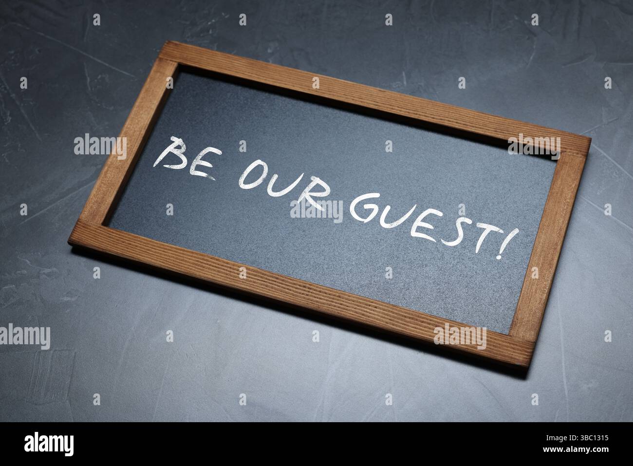 Piccola lavagna con la frase Be Our Guest su sfondo grigio scuro, vista dall'alto Foto Stock