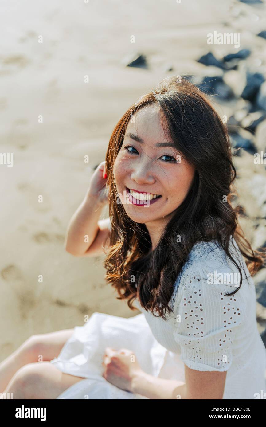 Una donna con i capelli lunghi è seduta sulla spiaggia e sorride. Indossa un vestito bianco e i capelli vengono tirati indietro Foto Stock