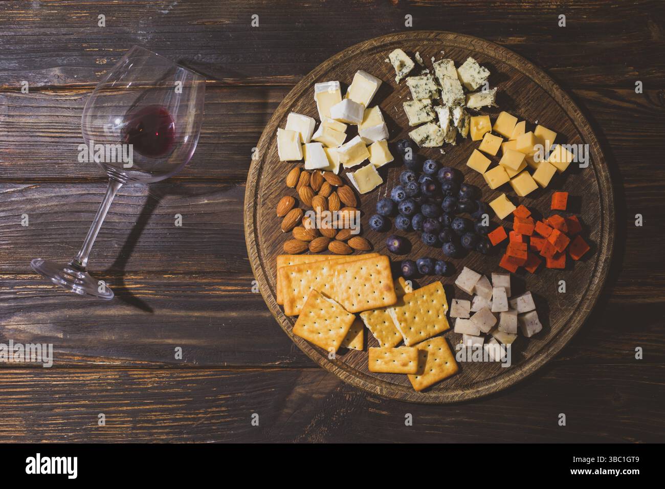 Il formaggio di diversi tipi e altri spuntini sono disposti su un vassoio di legno Accanto al tavolo di legno si trova un bicchiere con una goccia di vino rosso Foto Stock