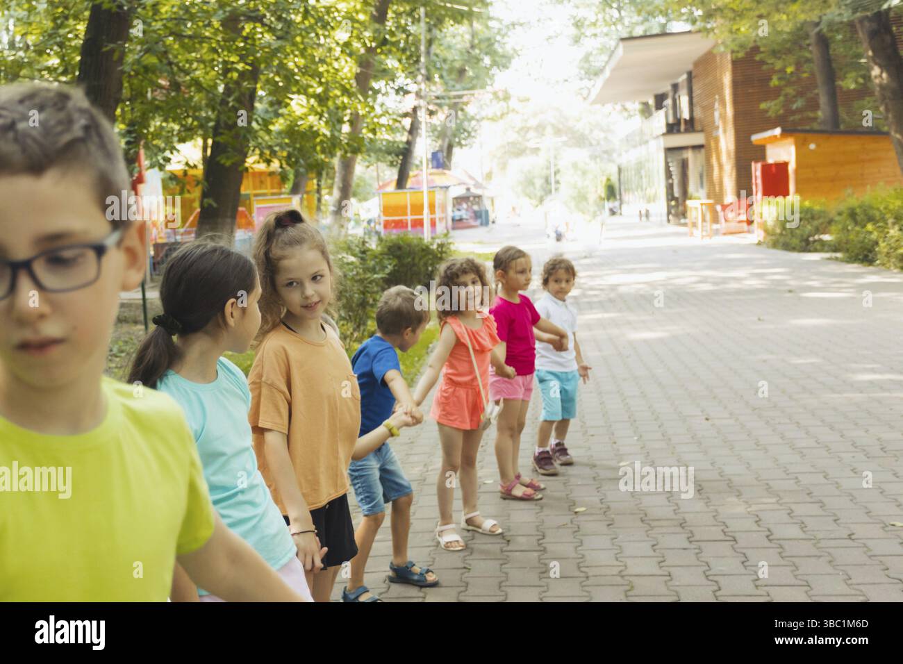 Bambini in abiti colorati in fila, tenendosi per mano. Giochi attivi all'aperto per bambini in età prescolare. Campo estivo nel parco cittadino Foto Stock