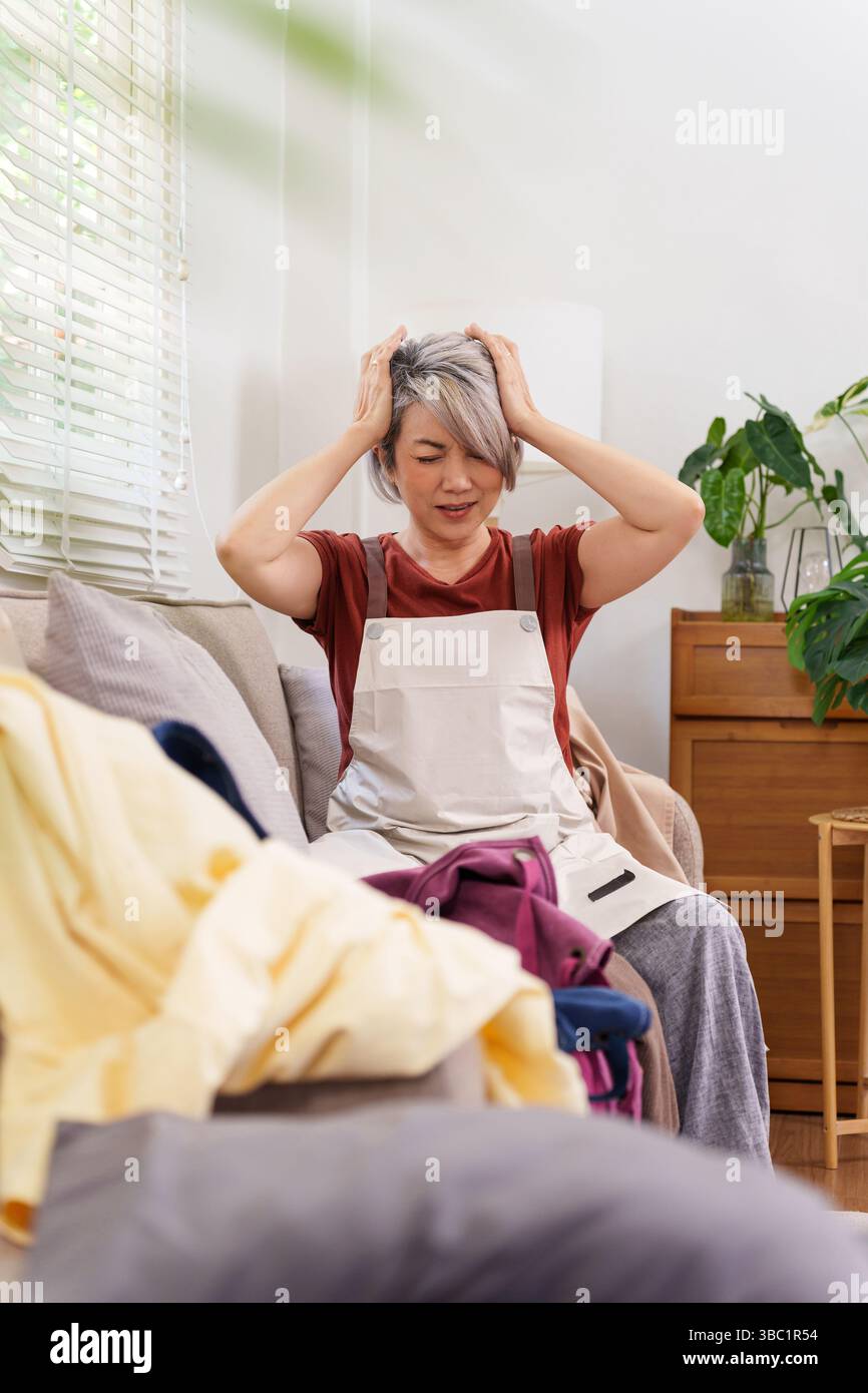 Donna adulta che indossa il grembiule che soffre di stress con un mucchio di bucato. Le faccende domestiche possono essere travolgenti. Concentrarsi sul burnout e la salute mentale associati W Foto Stock