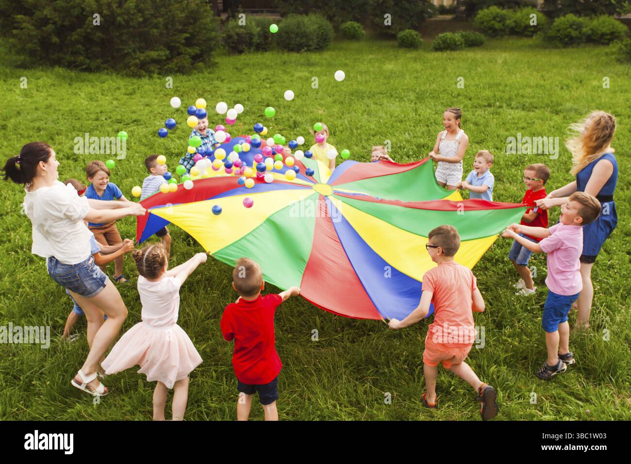 I bambini attivi che sventolano il paracadute multicolor grande per buttare fuori le palle. Giochi di gruppo per rafforzare il rapporto tra i bambini. Parco estivo con gras verde Foto Stock