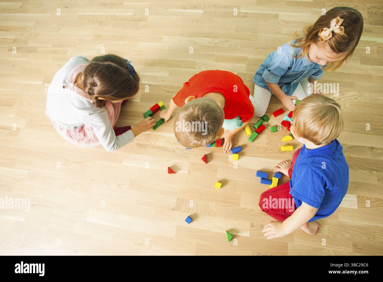 Quattro bambini giocano su un pavimento di legno con mattoni di legno colorati. Foto dall'alto dei bambini nelle torri giocattolo dell'asilo. Concetto di gioco libero Foto Stock