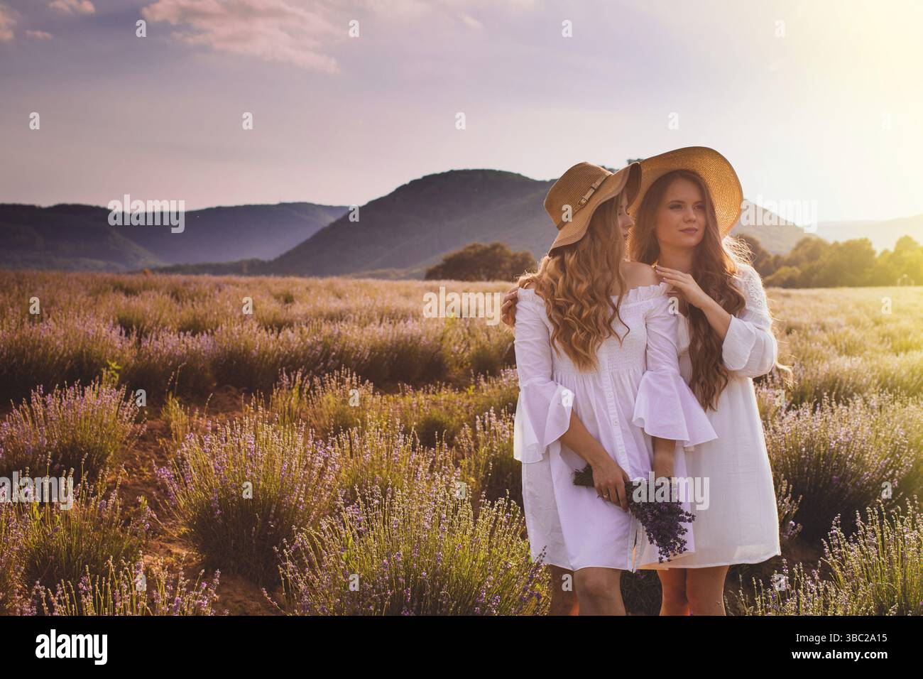 Due adorabili giovani donne, sorelle in corti abiti bianchi, con lunghi capelli sciolti, in piedi vicini l'uno all'altro mentre il tramonto in campo lavanda, nel retro l Foto Stock