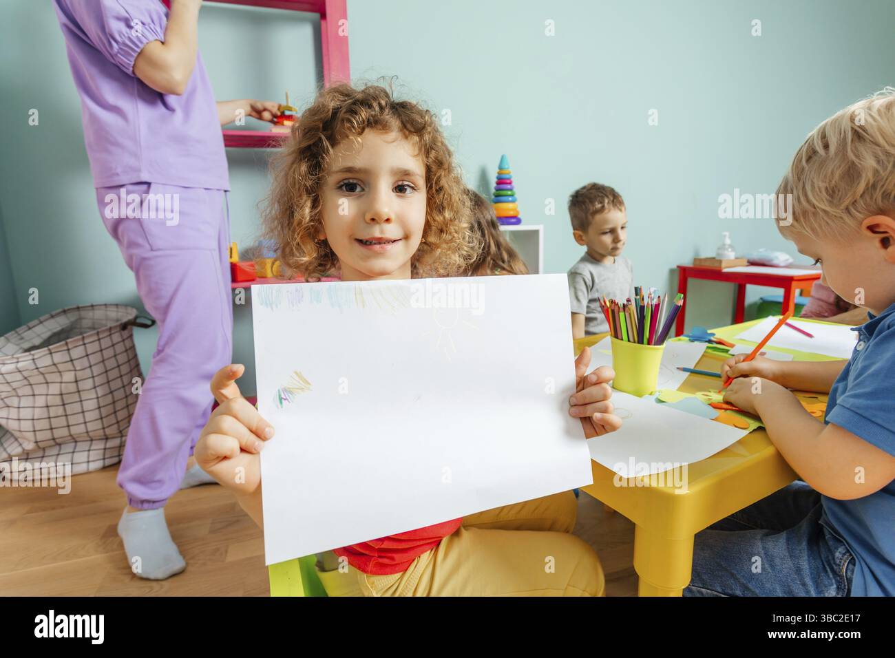 Ritratto di una bella ragazza riccia seduta, con un grande foglio di carta, che mostra il suo disegno alla fotocamera. Lezione d'arte all'asilo o all'asilo. Sentire Foto Stock