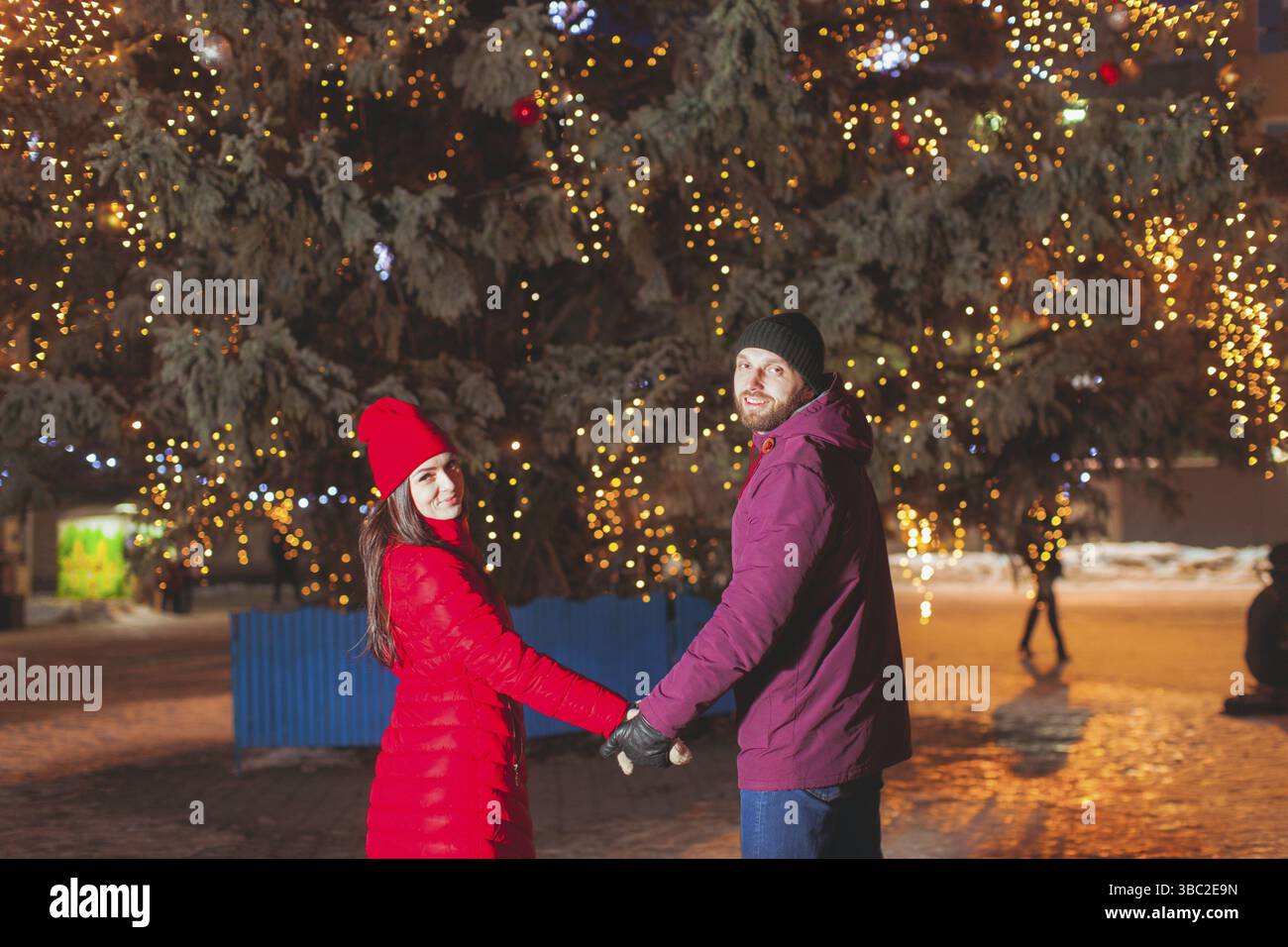 Ritratto di giovane coppia attraente posa vicino città albero di Natale decorato con luci. Sposi che viaggiano durante le vacanze invernali, indossando un caldo stile Foto Stock