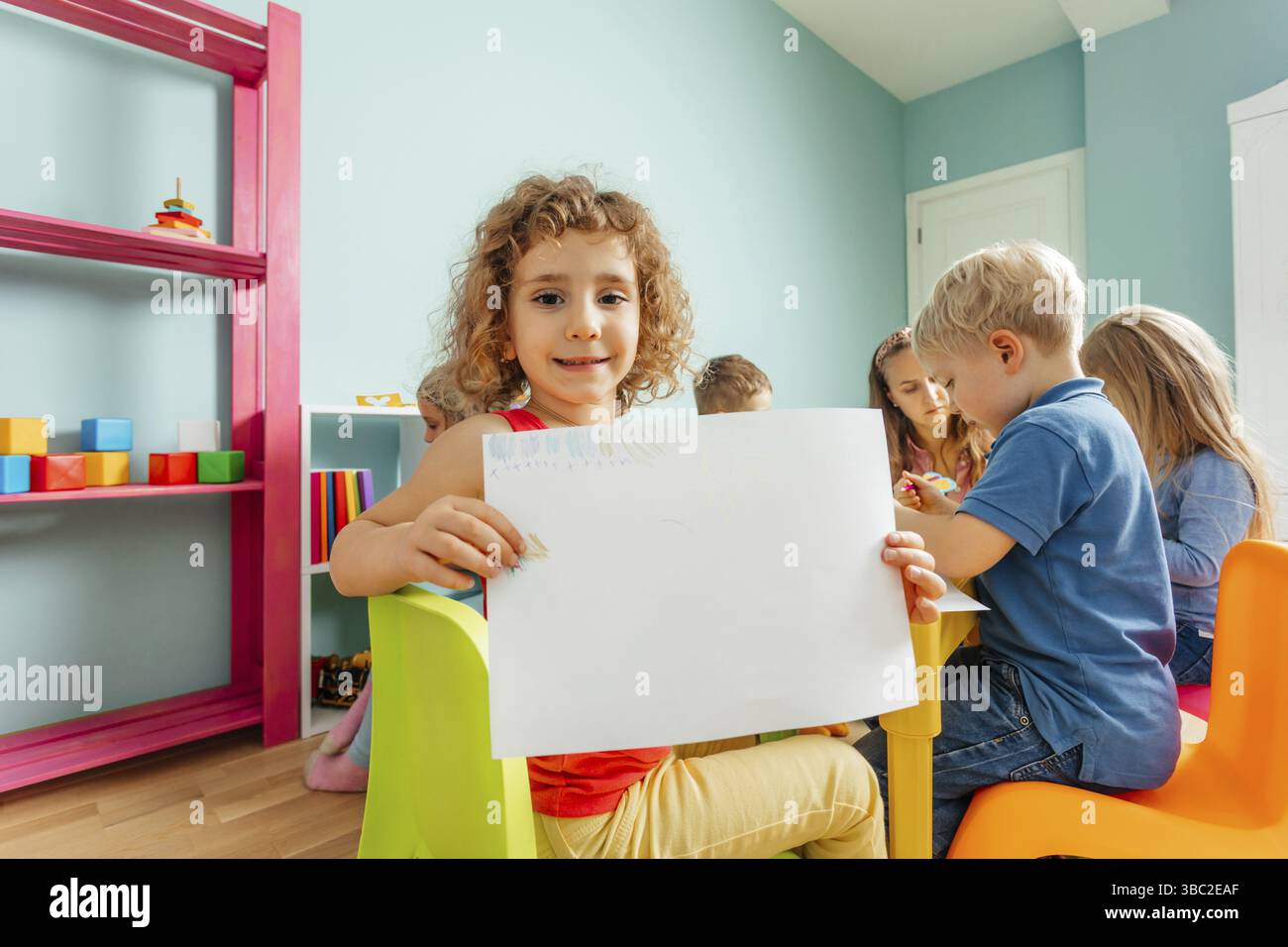 Ritratto di una bella ragazza riccia seduta, con un grande foglio di carta, che mostra il suo disegno alla fotocamera. Lezione d'arte all'asilo o all'asilo. Sentire Foto Stock