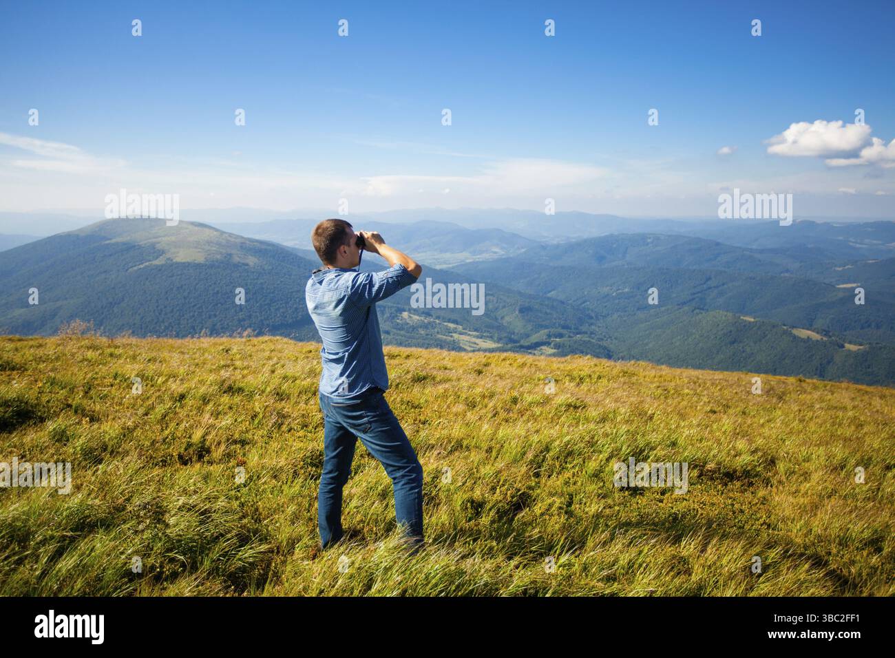Il giovane che indossa una camicia blu e jeans che guarda il binocolo e che gode di un paesaggio di montagna Foto Stock