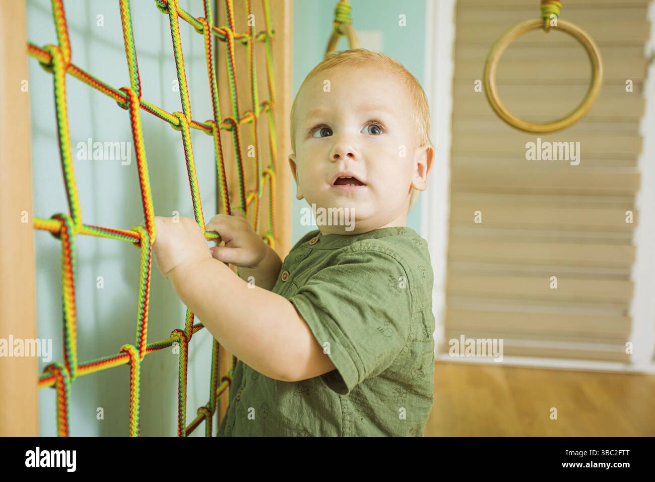 Ritratto di un bambino biondo che si arrampica sulla rete in una palestra di legno. Crescere i bambini organizzando lo spazio per l'attività fisica. Concetto di hobby salutare Foto Stock Ritratto di un bambino biondo che si arrampica sulla rete in una palestra di legno. Crescere i bambini organizzando lo spazio per l'attività fisica. Concetto di hobby salutare Foto Stock