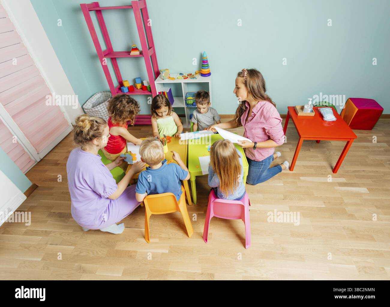 Lezione d'arte all'asilo, vista dall'alto. Bambini adorabili che incollano e tagliano la carta su tavoli di plastica colorati. Concetto di sviluppo per bambini Foto Stock
