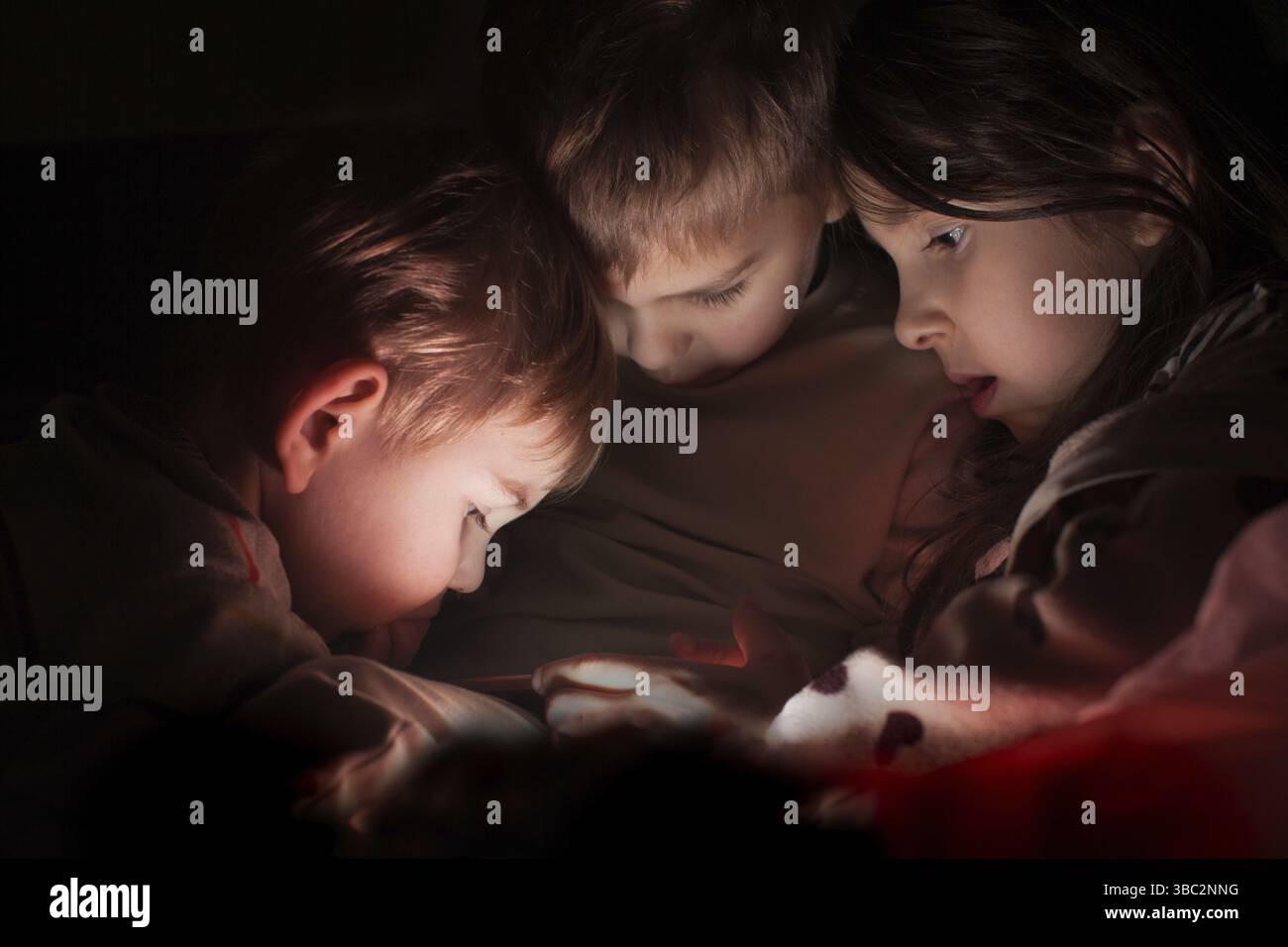 Bambini che giocano con uno smartphone in condizioni di luce oscura, con effetti negativi sugli occhi del bambino. Famiglia prima di dormire Foto Stock