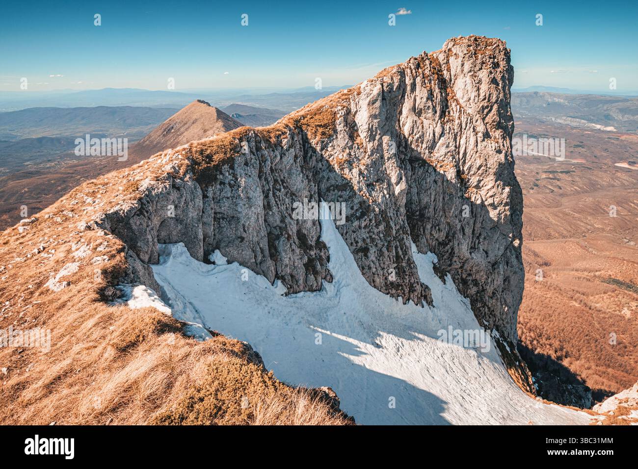 La neve, aggrappata alle ripide pendici di un'aspra cima di montagna, crea un contrasto drammatico con la vallata sottostante Foto Stock