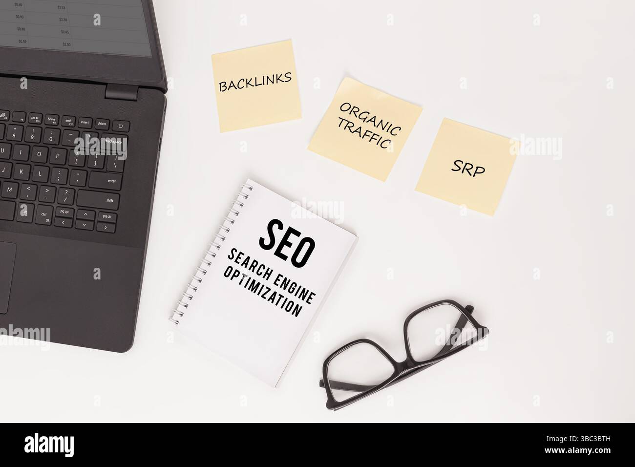 "Disposizione piatta di un laptop, occhiali, notebook etichettato "SEO Search Engine Optimization" e "BACKLINKS", "TRAFFICO ORGANICO" e "SRP"." Foto Stock