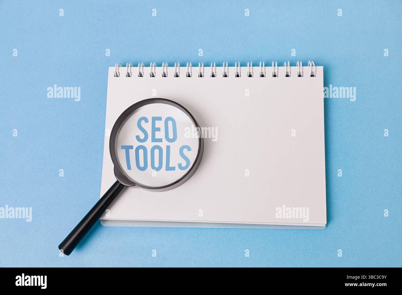 Una lente di ingrandimento con le parole "SEO TOOLS" ingrandite sotto poggia su un blocco note vuoto su uno sfondo azzurro. Foto Stock
