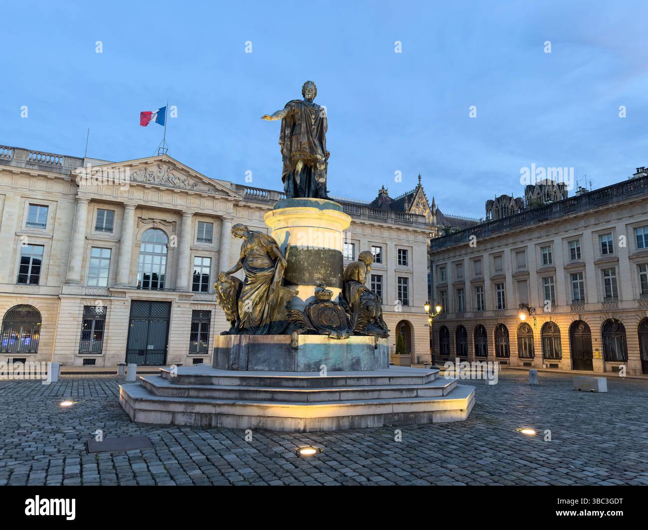 La statua in bronzo di Jean-Baptiste Colbert si erge in una piazza di Reims, illuminata da luci soffuse sullo sfondo del cielo serale. Foto Stock
