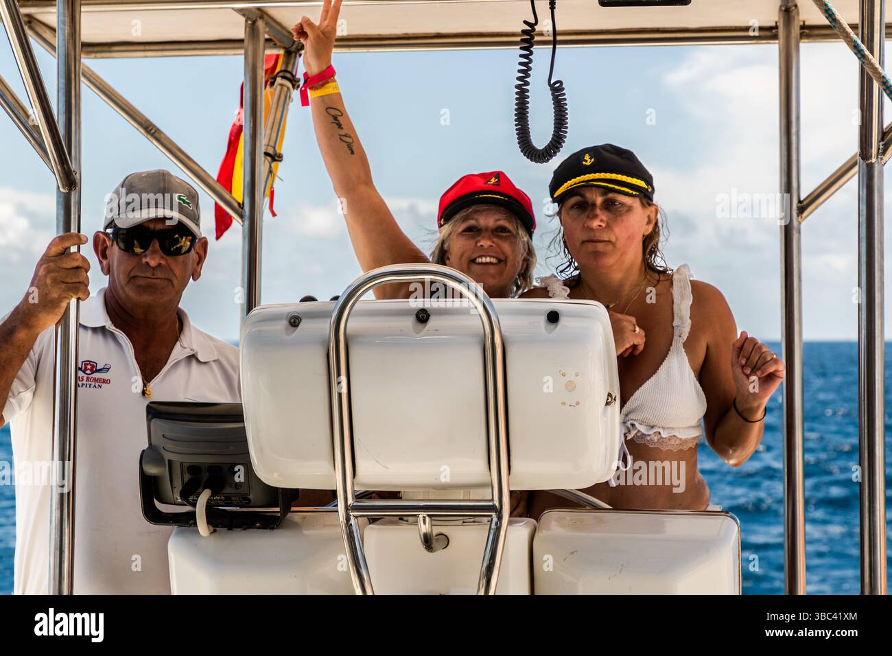 Il capitano della compagnia di navigazione Líneas Romero guida un catamarano al largo della costa di Lanzarote, mentre due passeggeri con un berretto da capitano e un top bikini posano allegramente. Líneas Romero opera escursioni e servizi di traghetto tra Lanzarote, Fuerteventura e La Graciosa. Costa de Papagayo, Isole Canarie, Spagna. Foto Stock
