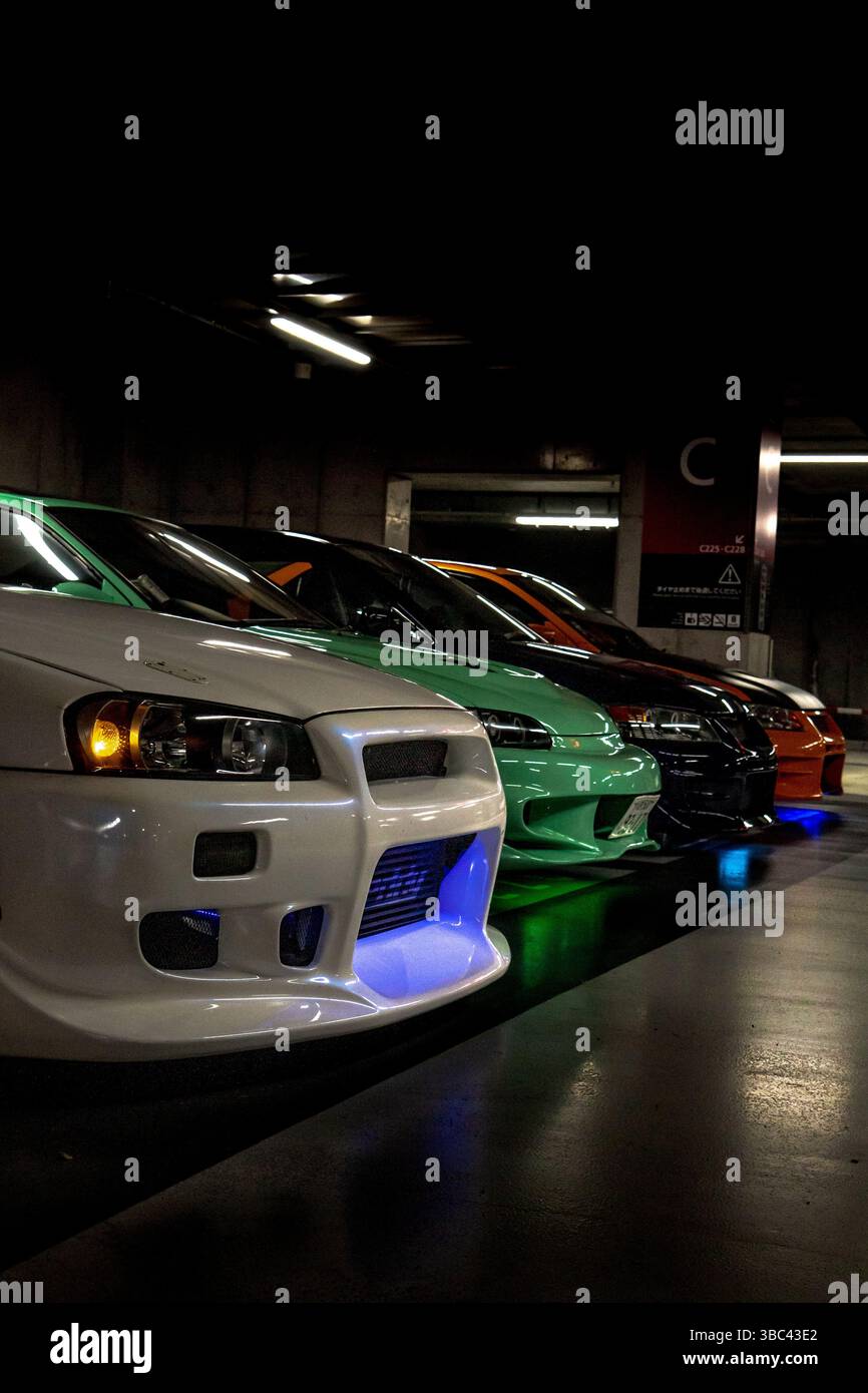 Una gamma di auto sportive giapponesi modificate in un incontro sotterraneo di Tokyo, illuminate da colorate luci al neon Foto Stock