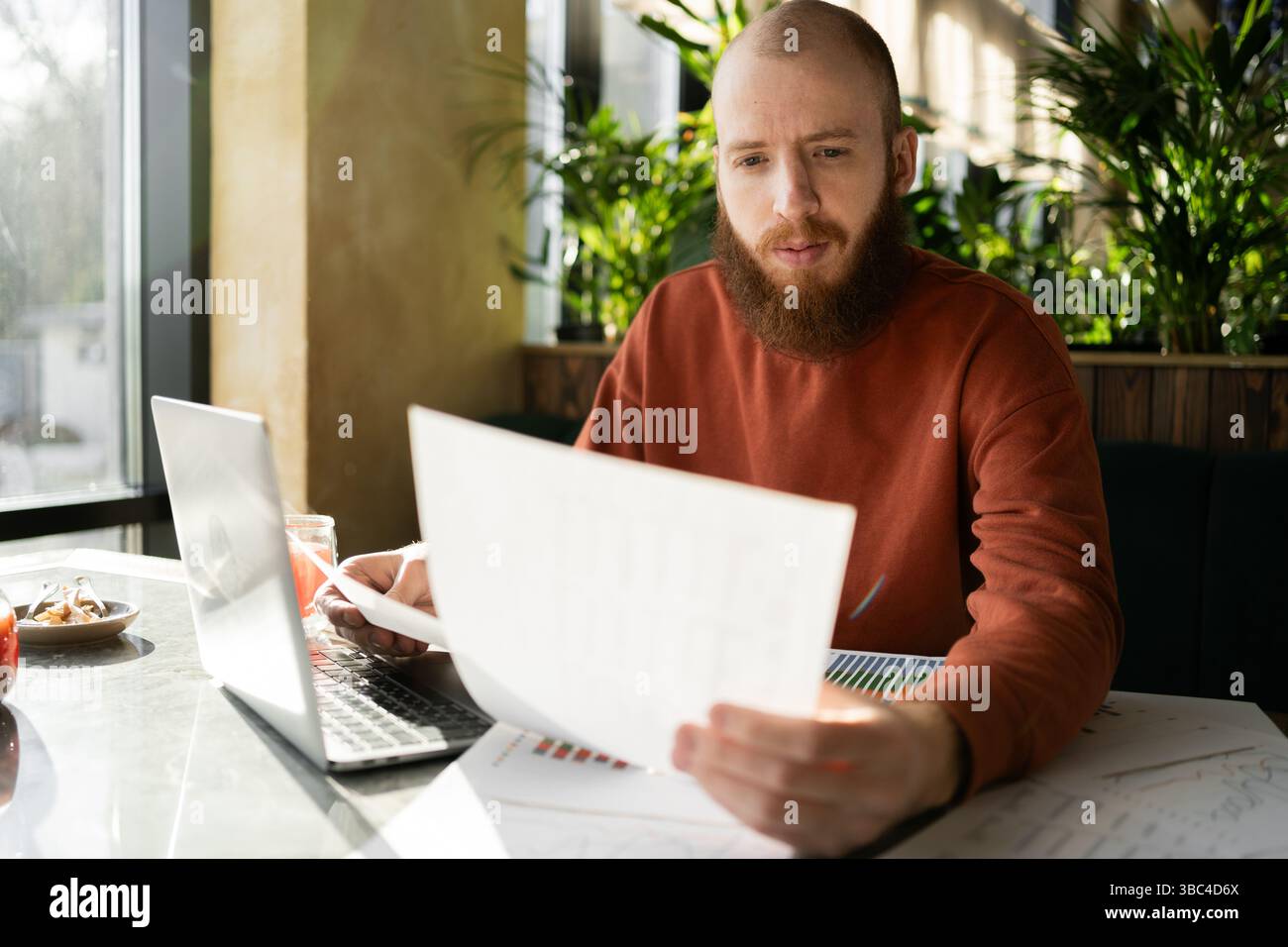 imprenditorialità. Bearded bell'uomo che legge un documento seduto con un computer portatile in un bar vicino alla finestra. Moderna startup e pianificazione aziendale Foto Stock