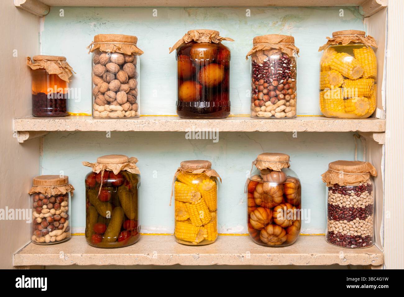 Vari cibi conservati, tra cui noci, mais, fagioli e sottaceti, conservati in vasetti di vetro con coperchi di carta su scaffalature rustiche. Foto Stock