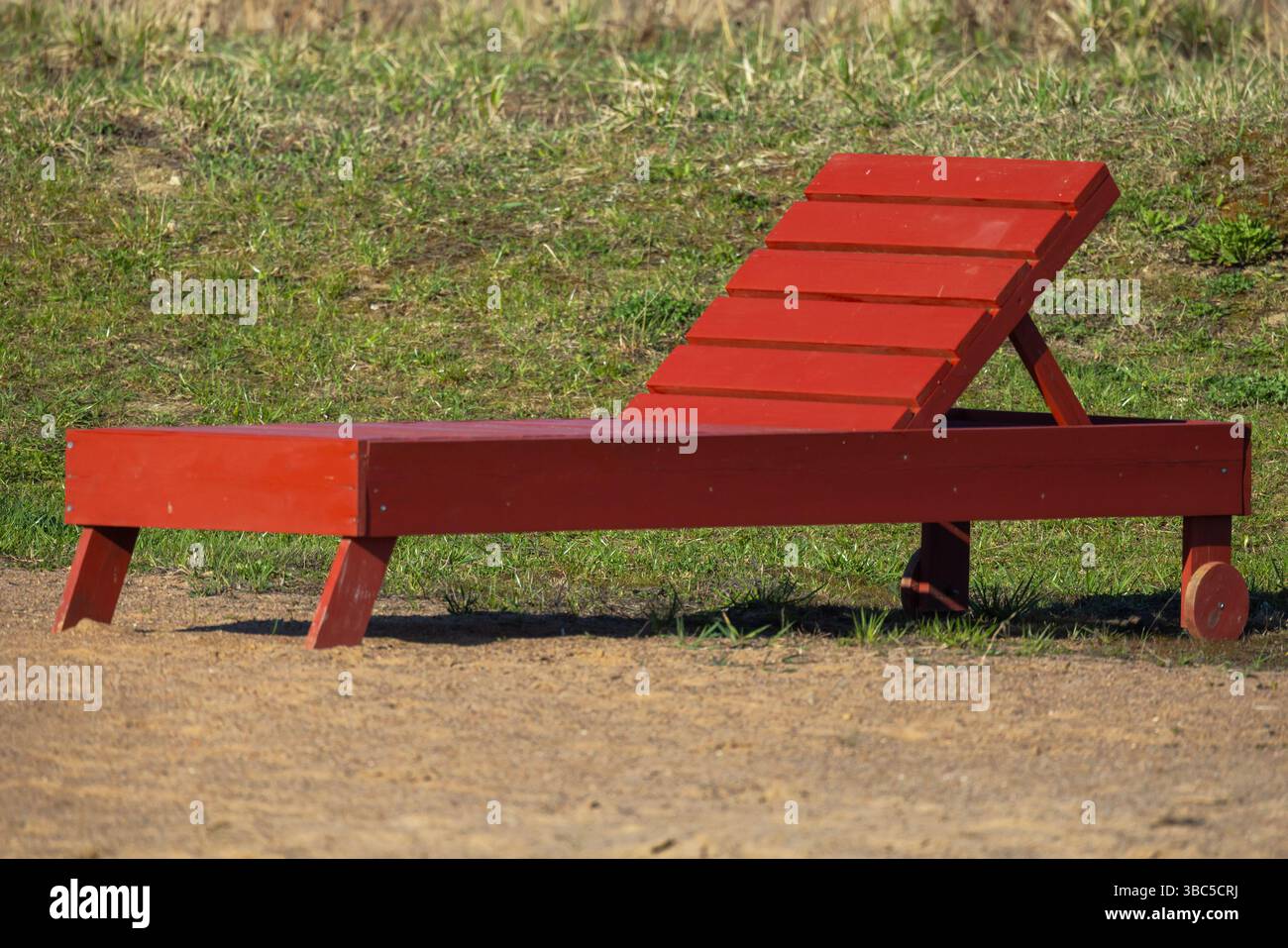 Ambiente esterno caratterizzato da una sedia da spiaggia rossa in legno situata su una superficie sabbiosa con un ambiente erboso. La sedia è elegantemente realizzata, progettata per Foto Stock
