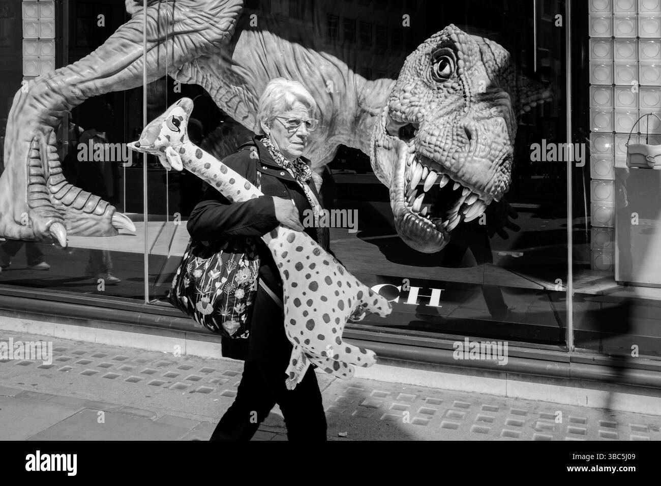 La donna che porta una giraffa giocattolo morbido passa davanti alla grande figura di Tyrannosaurus Rex nella vetrina del negozio. Regent Street, Londra, Regno Unito. Foto Stock