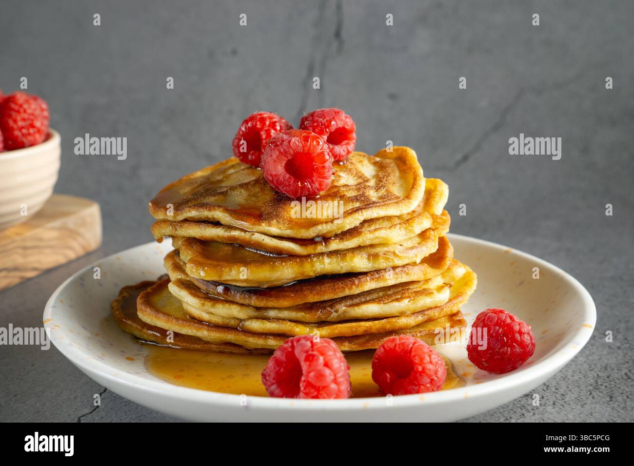 primo piano di una pila di frittelle di banana dorate su un piatto bianco, ricoperte di lamponi freschi e condite con sciroppo Foto Stock