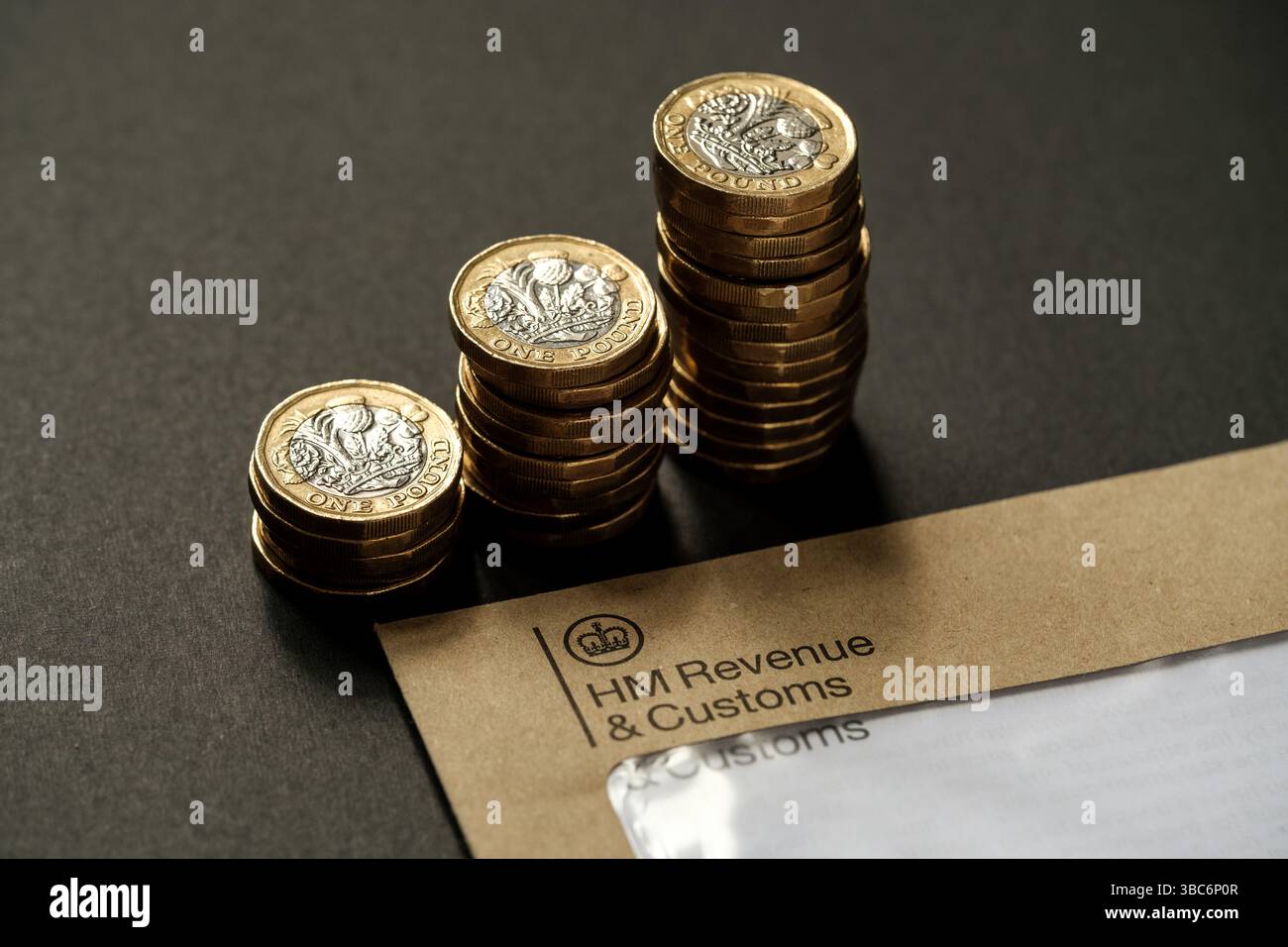 Monete inglesi impilate da una sterlina accanto alla busta HM Revenue & Customs su sfondo scuro, concetto di tassazione del Regno Unito e responsabilità finanziaria personale, Foto Stock