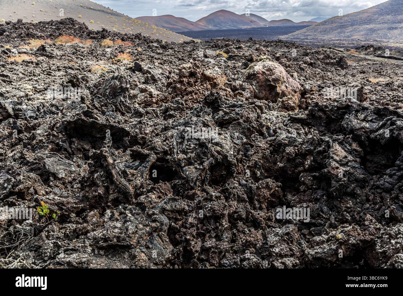 Un campo di lava si estende davanti a diversi coni vulcanici del Parque Natural de los Volcanes vicino a Mancha Blanca a Lanzarote, Isole Canarie, Spagna. Erbe gialle sparse crescono tra le rocce nere. Foto Stock