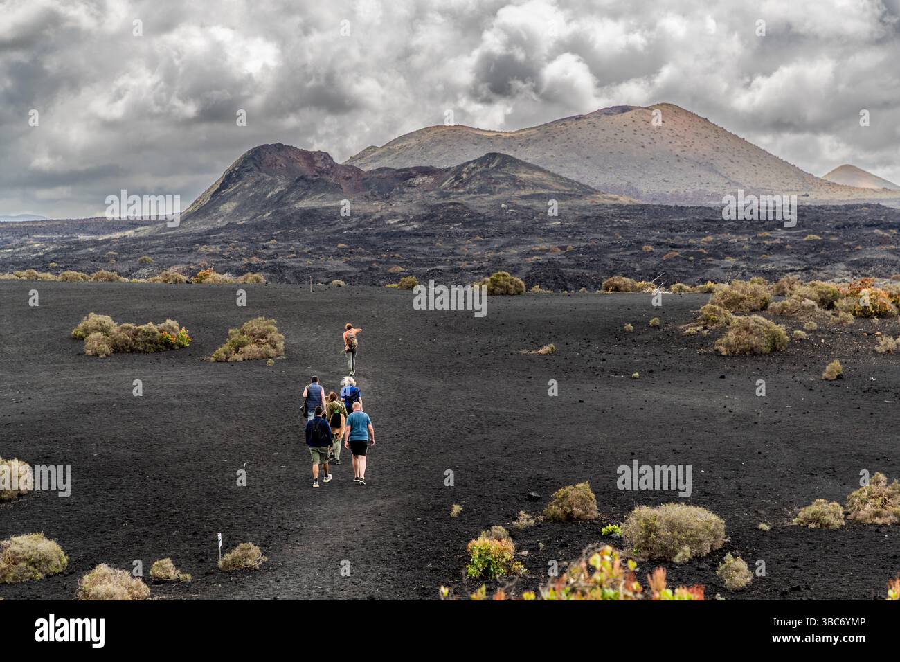 Un gruppo di escursionisti attraversa un campo di lapilli neri nell'area vulcanica vicino a Mancha Blanca a Lanzarote. Diversi coni vulcanici del Parque Natural de los Volcanes costeggiano l'orizzonte. Uno di essi ha una cima con sfumature rossastre. Arbusti secchi crescono sparsi tra le rocce. Isole Canarie, Spagna. Foto Stock