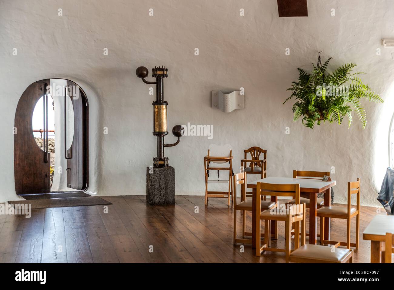 Uno strumento storico in ottone su un piedistallo di pietra si trova nell'interno pannellato in legno del Mirador del Río a Lanzarote, accanto a un portale ad arco e a una zona pranzo con tavoli e sedie. Isole Canarie, Spagna. Foto Stock