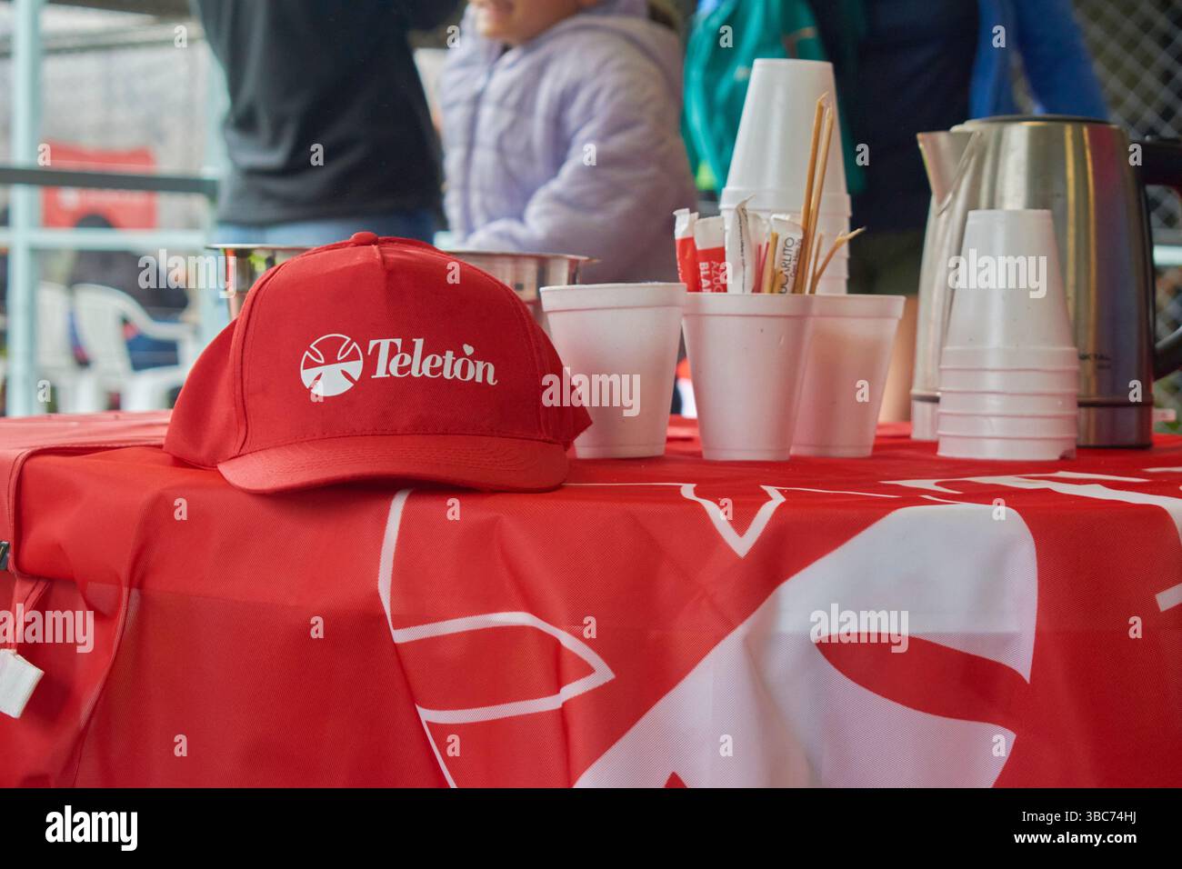 Un tavolo con una tovaglia rossa con il logo Teleton, su cui sono presenti un berretto, bicchieri, una teiera e sacchetti di zucchero. Sullo sfondo, gente fuori da fo Foto Stock