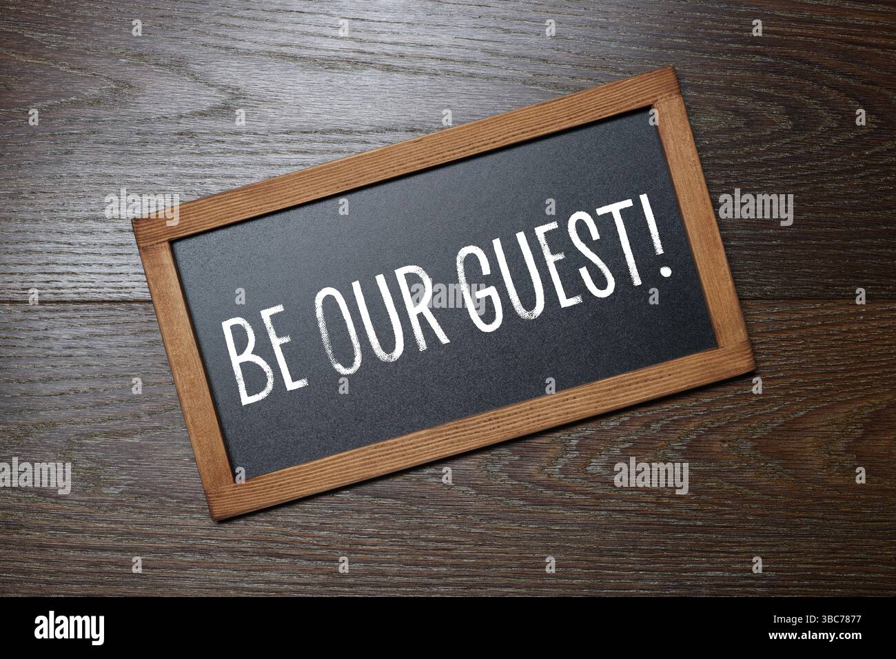 Piccola lavagna con frase Be Our Guest su sfondo in legno, vista dall'alto Foto Stock
