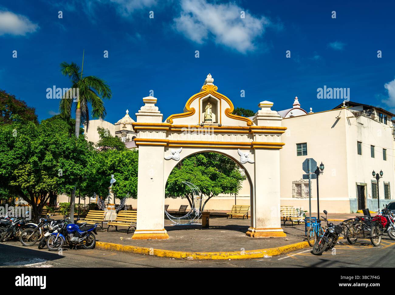 Arco ornamentale in stile coloniale si erge prominentemente nel Parque de la Merced nel centro di Leon, Nicaragua. Incorniciata da motociclette, palme e panchine, la struttura evoca il ricco patrimonio architettonico della città sotto un cielo azzurro Foto Stock